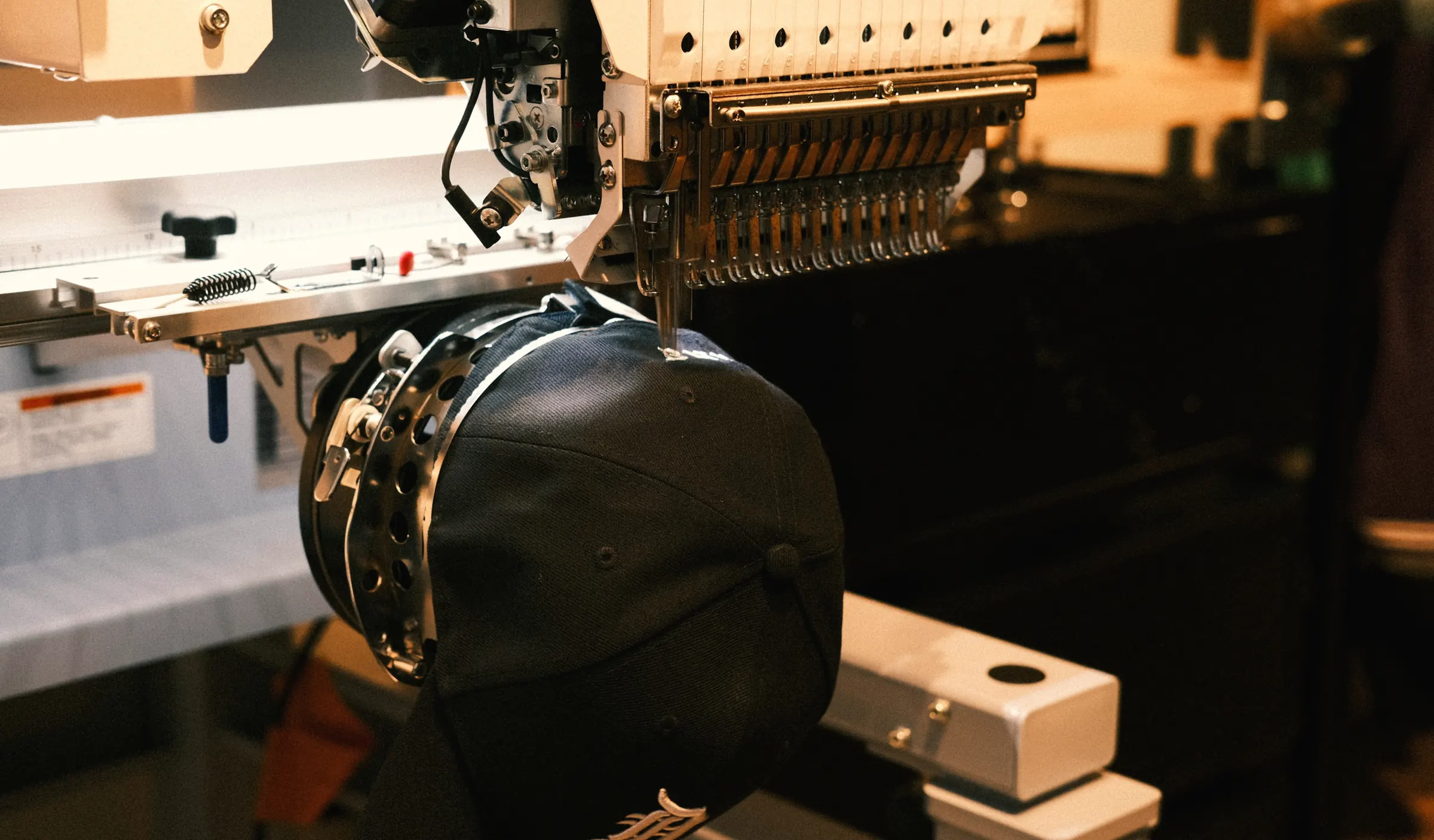 A cap clamped on a multi-needle embroidery machine, lit warmly in a dark workshop