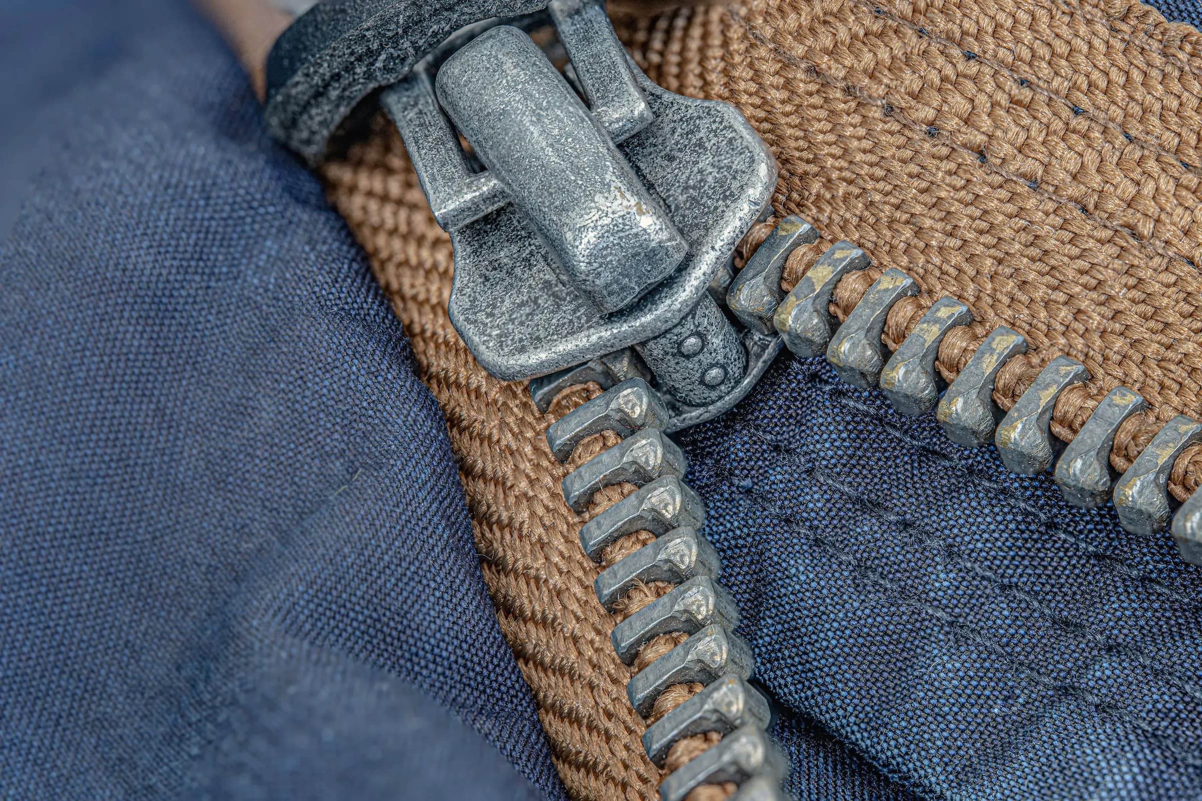 Macro of a chunky antique-nickel zipper and pull on navy canvas with tan webbing tape