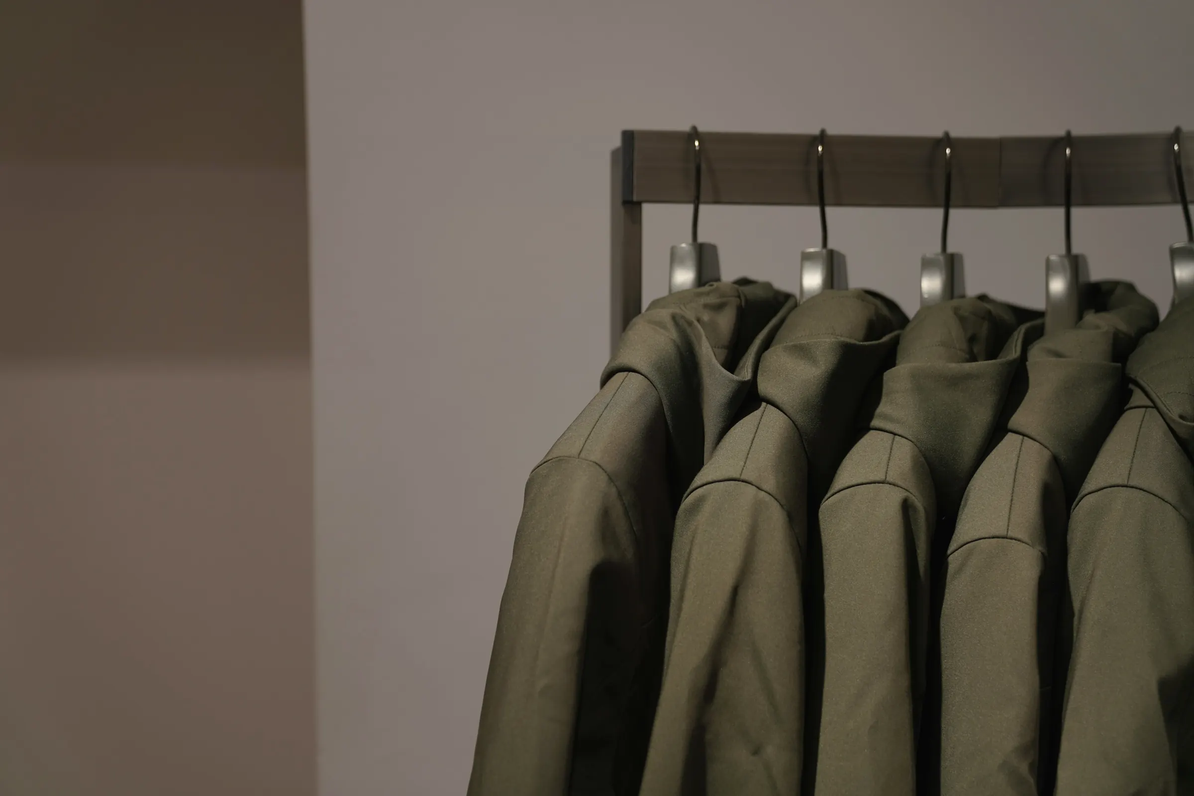 A row of olive-green cotton jackets hanging on a dark rail against a warm grey wall