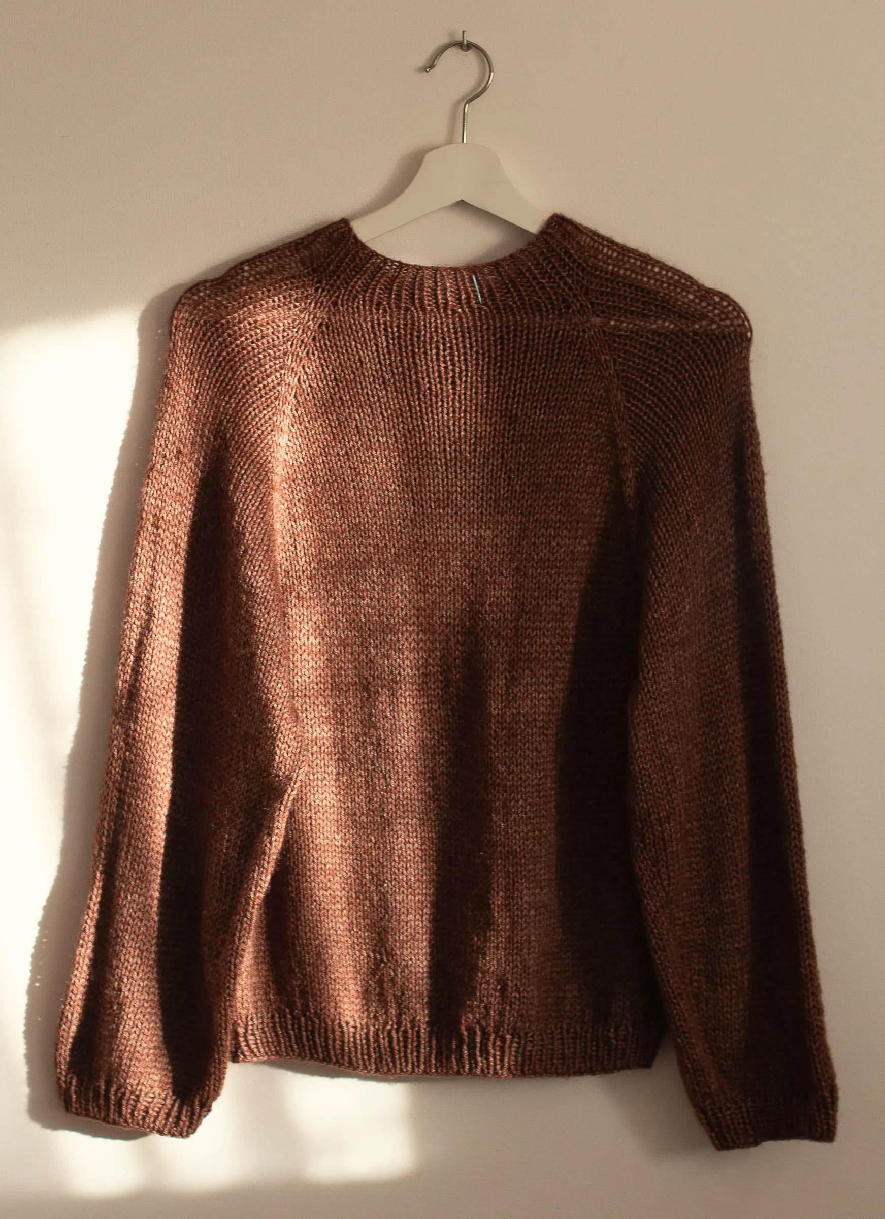 A chestnut-brown knit crewneck hanging on a white hanger in soft window light