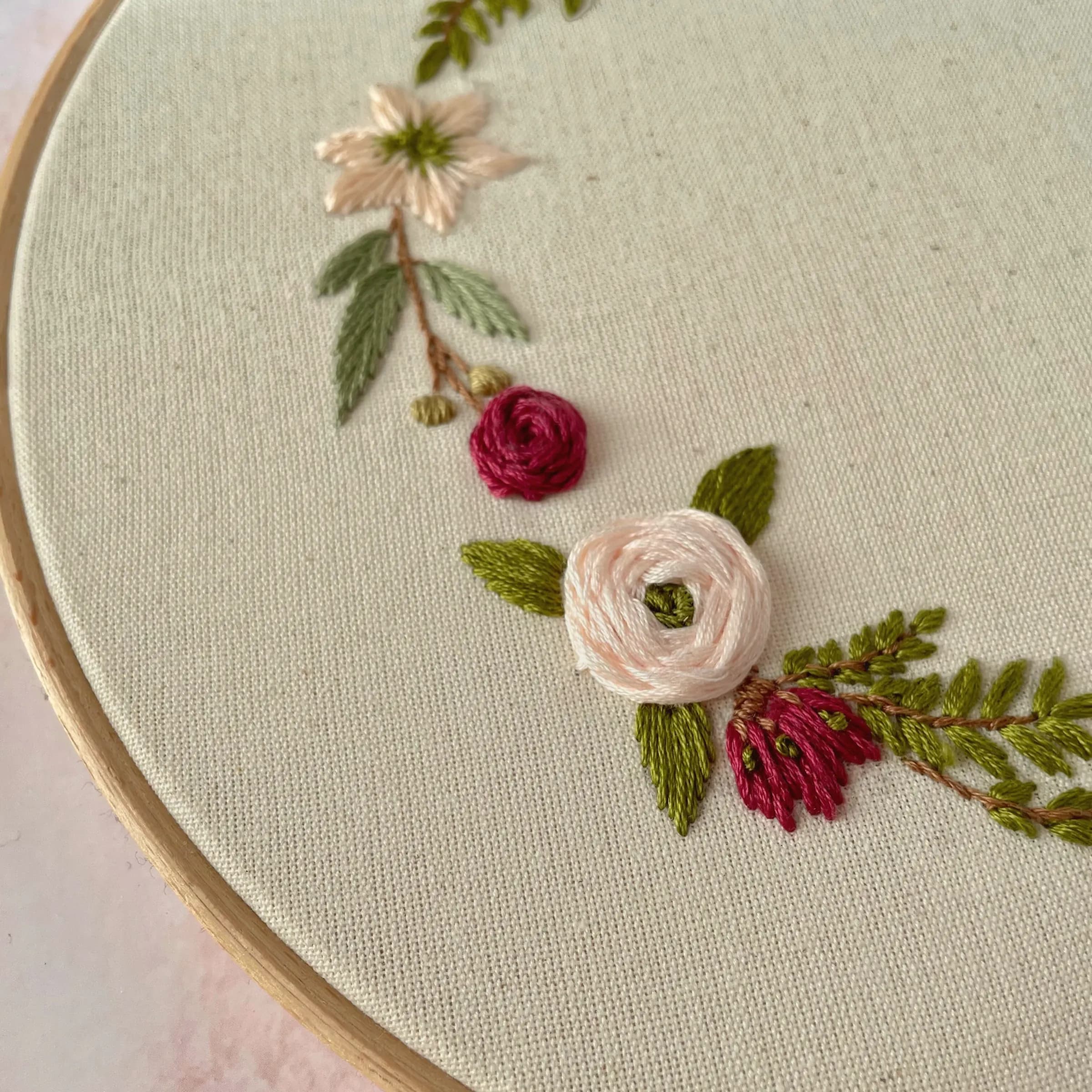 Close-up of hand embroidery in a wooden hoop: blush and crimson satin-stitch roses with green leaves on natural cotton