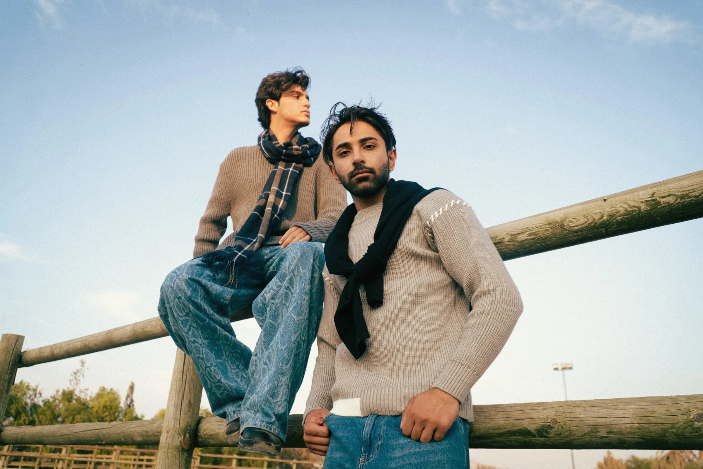Two young men on a weathered wooden fence, one in a ribbed knit with a plaid scarf, the other in an oatmeal sweater with a black knit tied over his shoulders
