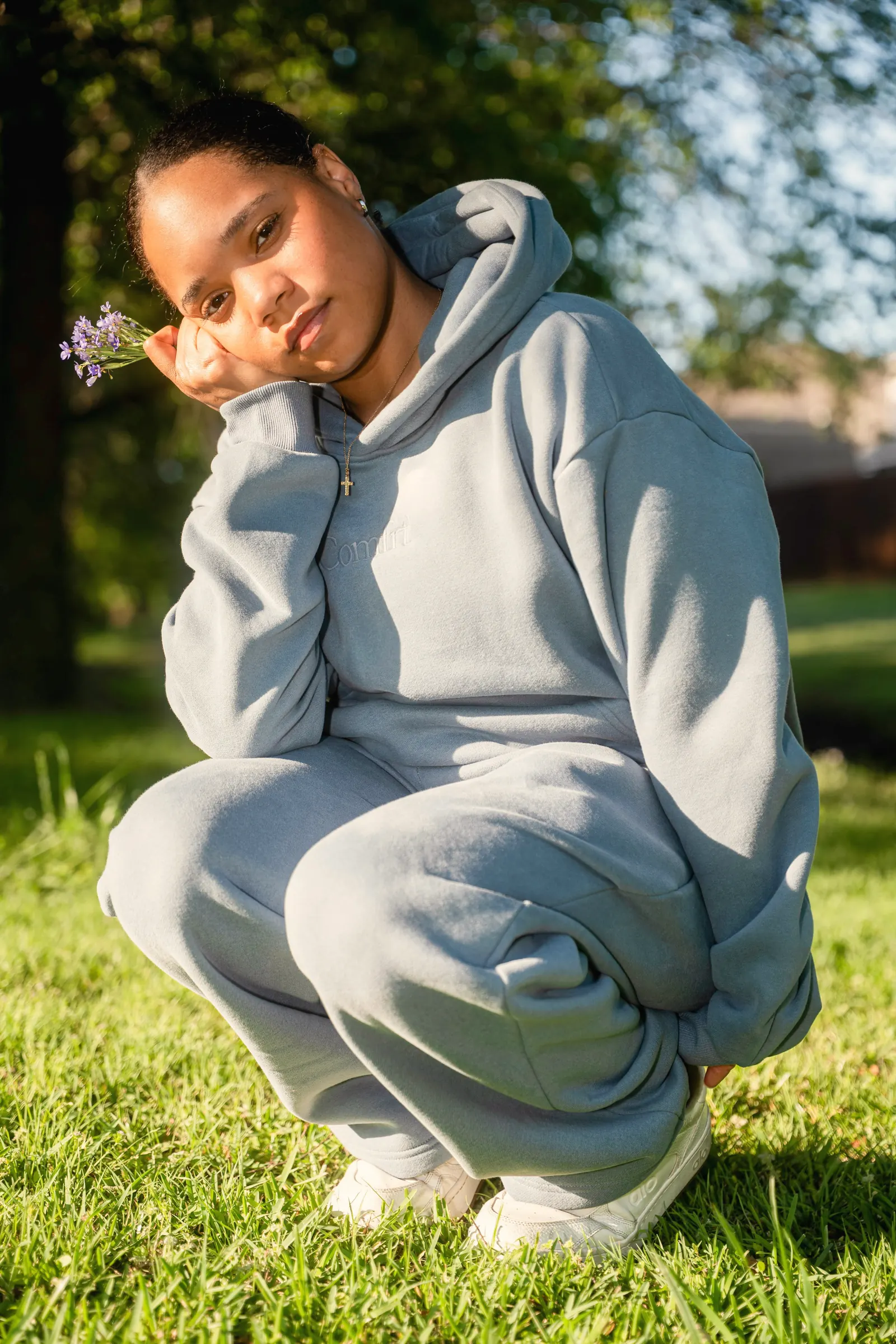 A young woman crouching on sunlit grass in a matching pale-blue fleece hoodie and sweatpants, holding a sprig of lavender